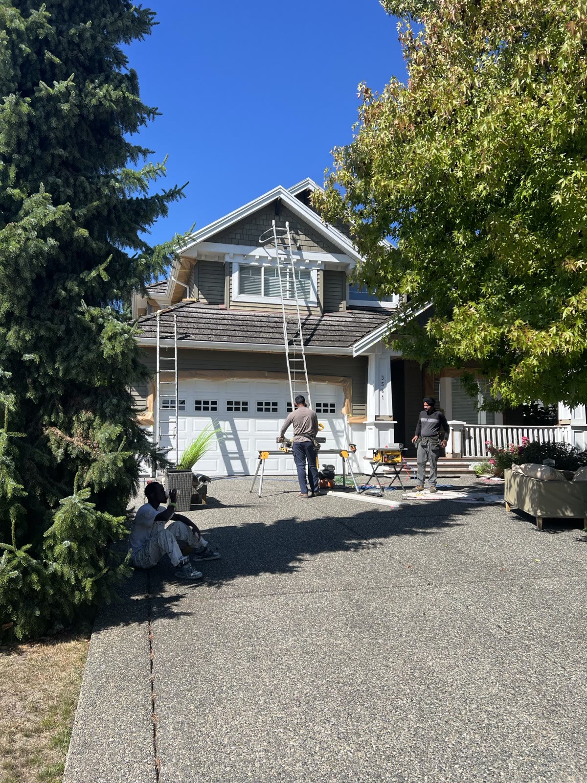 Dave's crew on the driveway of the same house — ladders up against the roofline, saw table and sprayer gear out